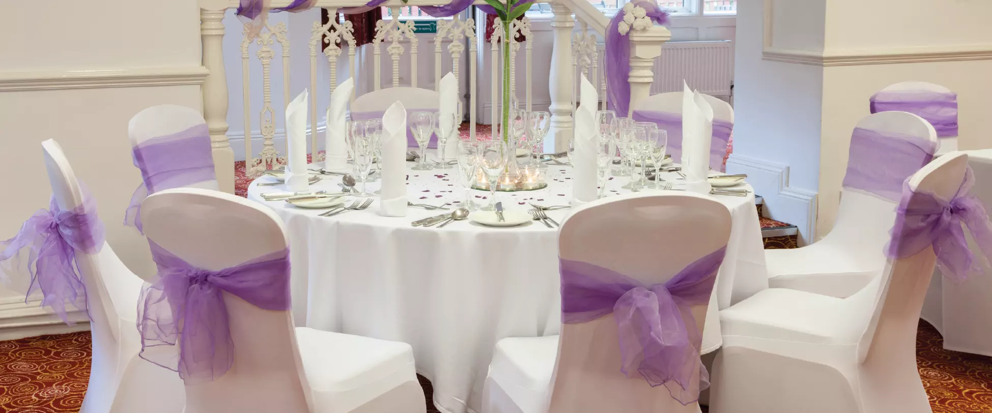 A table set up for a wedding with white table and chair covers. The chairs have purple ribbons and a tall centrepiece..