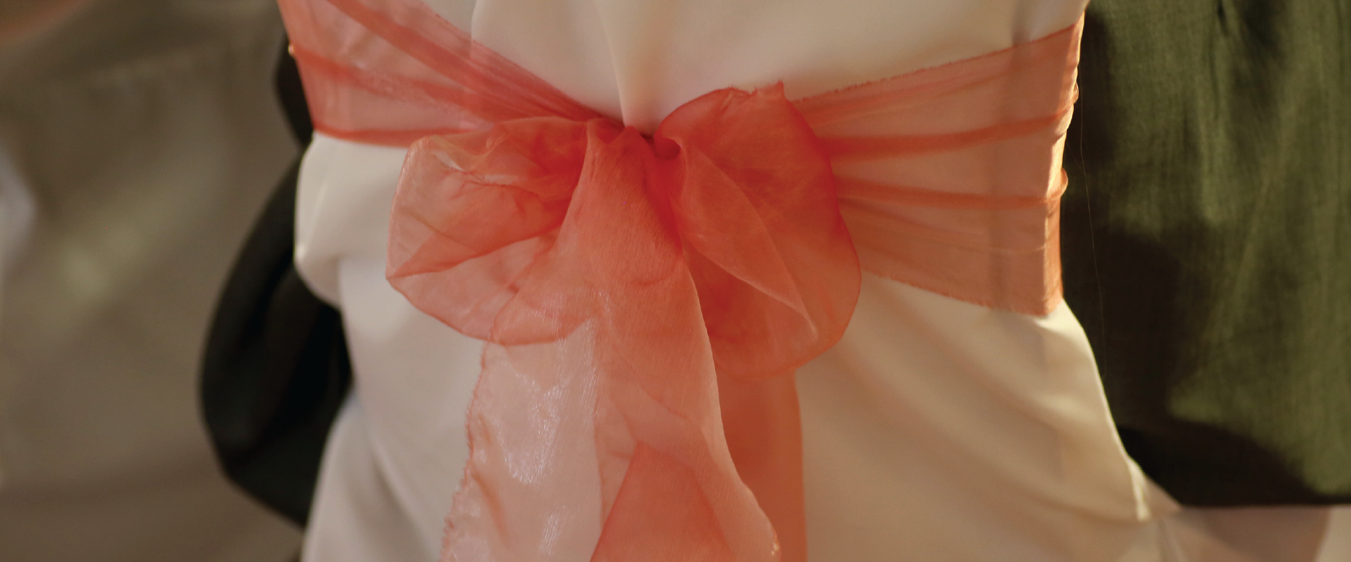The back of a chair dressed for a wedding with a white cover and peach ribbon..