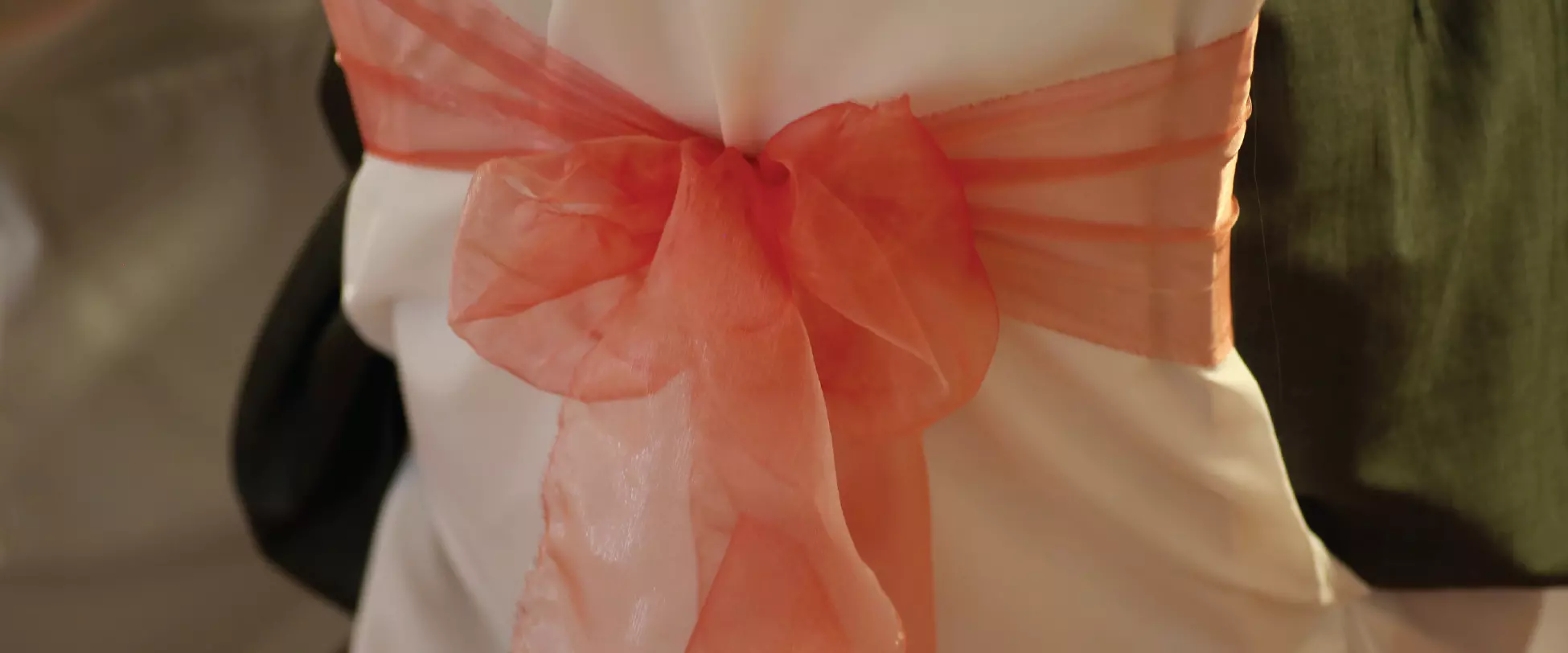 The back of a chair dressed for a wedding with a white cover and peach ribbon..