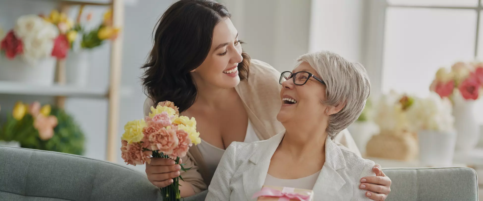 A woman with long brown hair smiling and embracing her mother, a woman with short grey hair and glasses. The first woman is hold a bouquet of pink and yellow flowers. There are an array of pink, yellow and white flowers in the background..